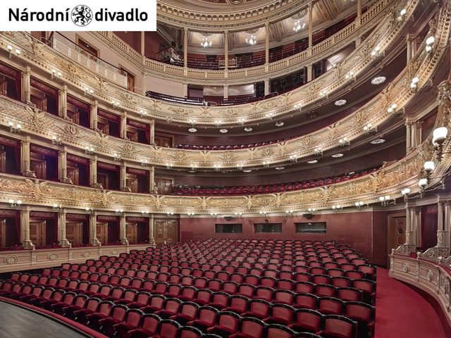 Binnenkant van een grote theaterzaal met rode stoelen en meerdere zitniveaus erboven. Het logo van het Nationaal Theater Praag in de linkerbovenhoek