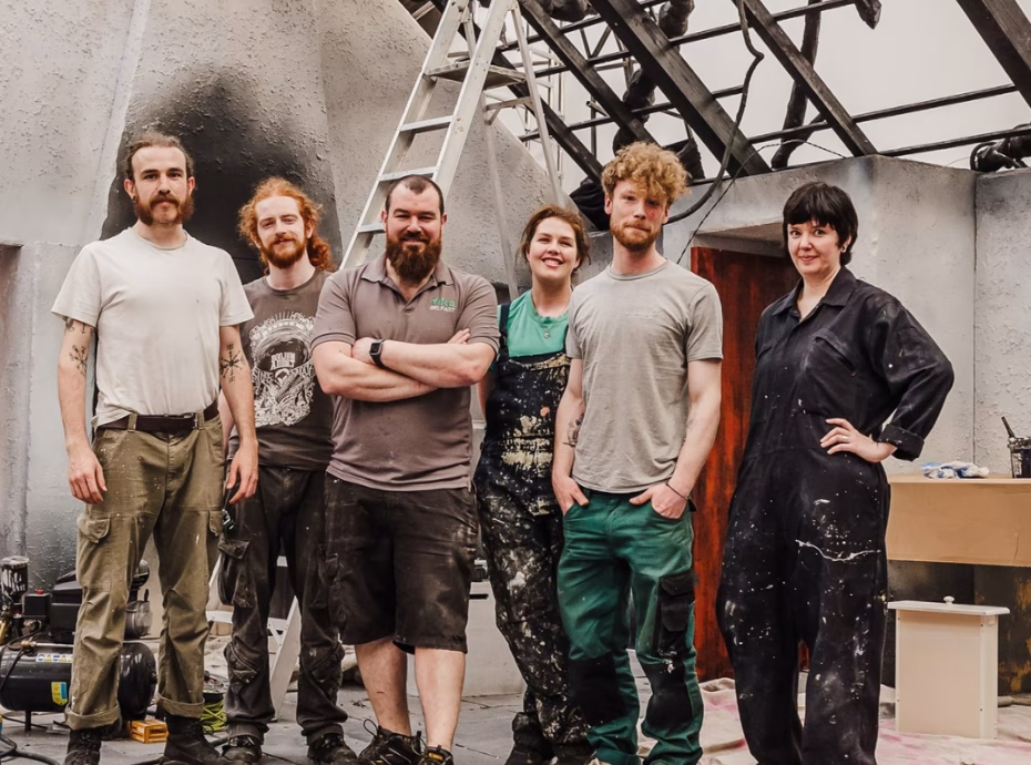 Binnenkant van een grote theaterzaal met rode stoelen en meerdere zitniveaus erboven. Het logo van het Nationaal Theater Praag in de linkerbovenhoek